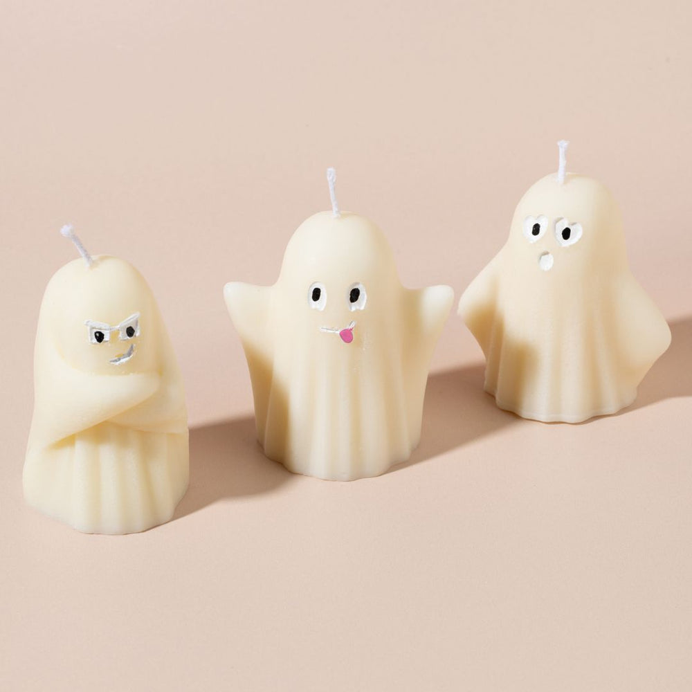 Three ghost-shaped candles with facial expressions on a beige background